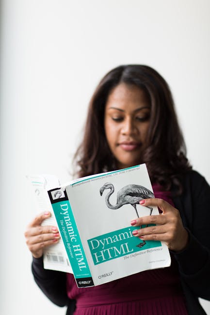 A woman with dark hair, dressed in a black blazer and maroon top, is standing indoors holding an open book titled 'Dynamic HTML' by O'Reilly, which features a detailed illustration of a flamingo on th