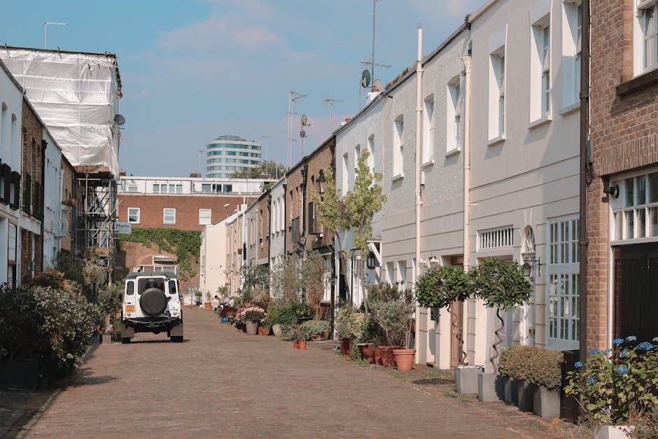 A narrow brick alleyway used for home relocation or furniture transport in Fulham Road Chelsea, with a paved pathway flanked by high brick walls on both sides. On the left, there are foliage and small