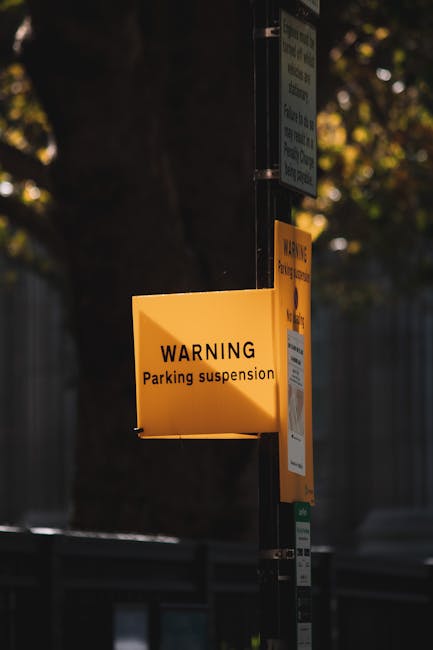 A yellow warning sign attached to a black metal pole indicating parking suspension, positioned outdoors with trees and a fence in the background. Below the warning sign, there are other permit or info